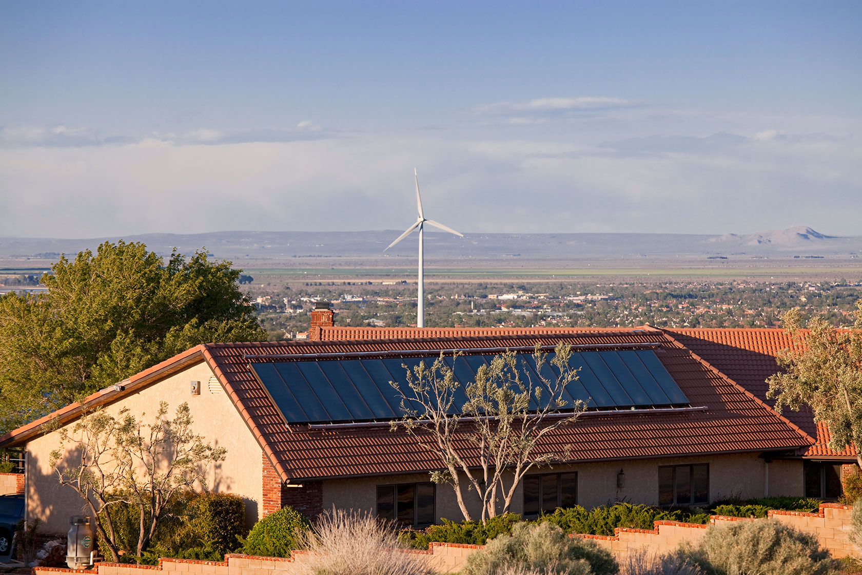 Photo shows a house with solar panels on its roof and a wind turbine in the background.