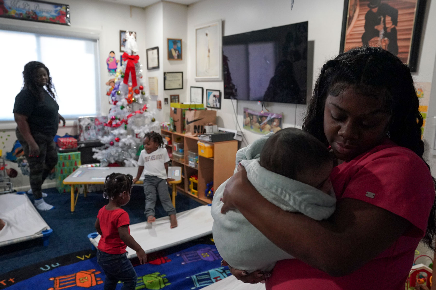 Photo shows a woman in the foreground holding a baby with another teacher and toddlers in the background, in a room filled with toys.