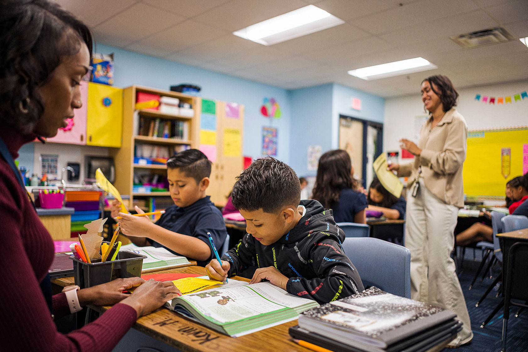 Photo shows a woman in the foreground helping students working on a project with books on their desk, as a second woman, the teacher, looks on.