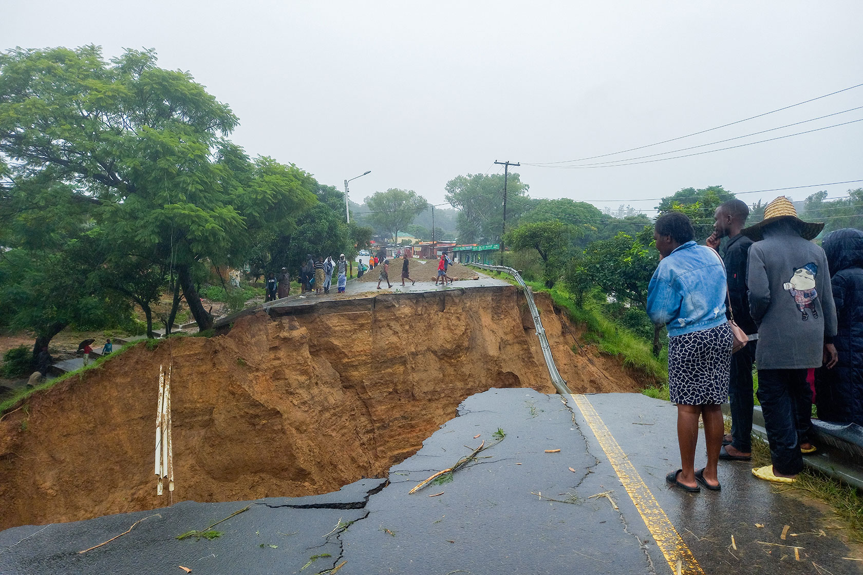 Photo shows people at the edge of a road town apart by flooding, with a giant crater in between the broken sides.