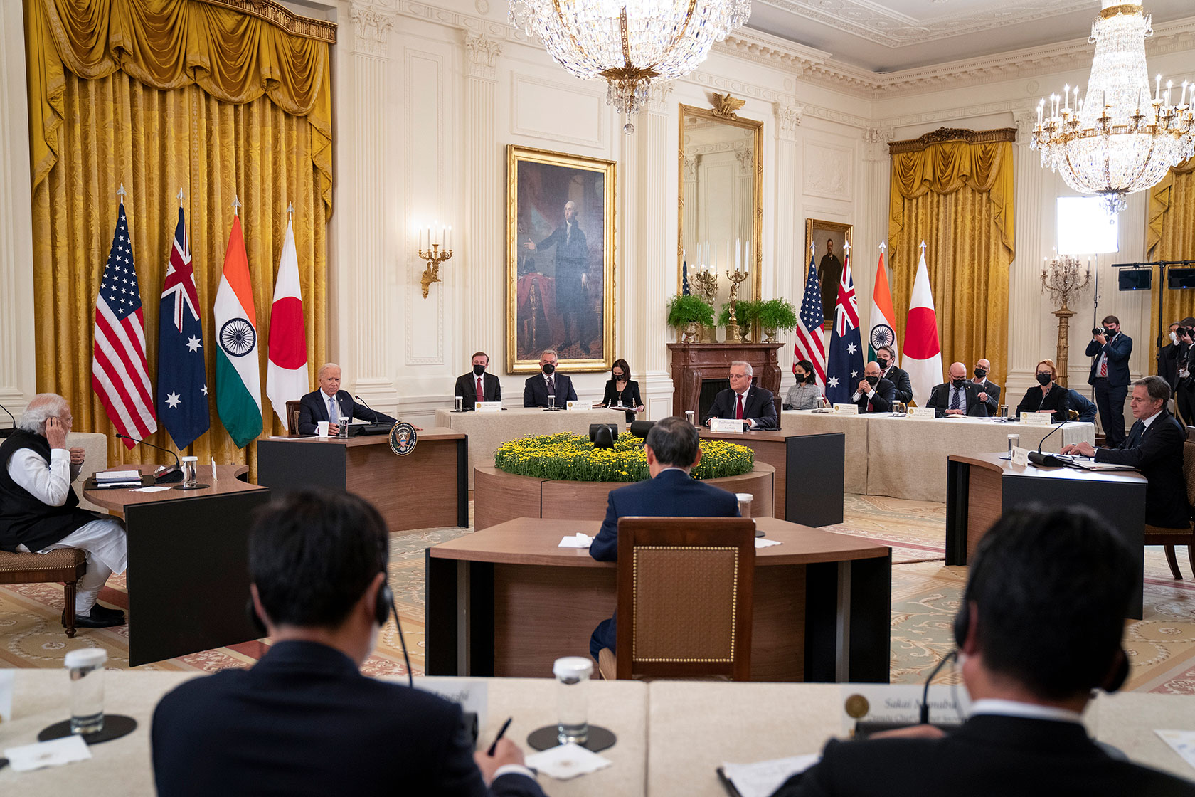 Photo shows Joe Biden, Narendra Modi, Scott Morrison, and Yoshihide Suga sitting at tables and facing each other, with other staff sitting around the perimeter, in a room at the White House.