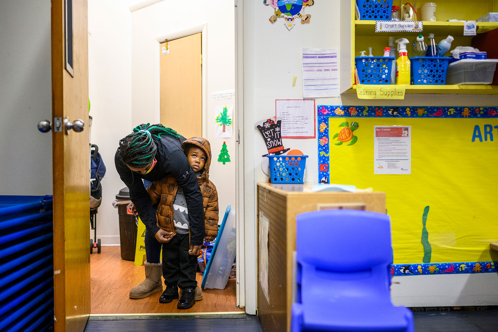 An early childhood educator zips up the coat of her 3-year-old student .