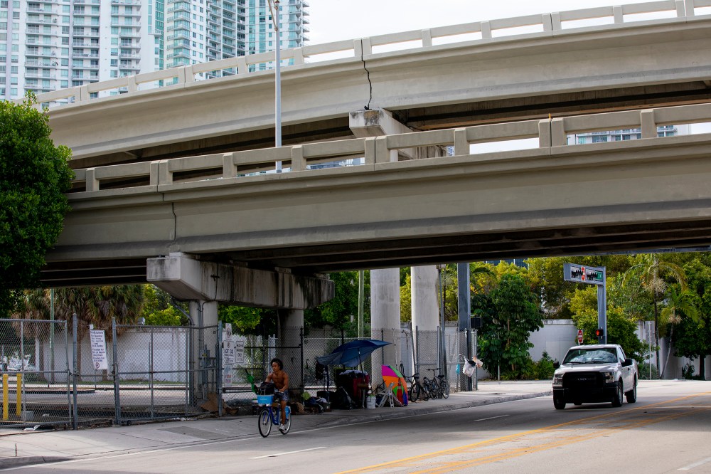 Makeshift living quarters underneath bridge alongside road with car and person on bicycle