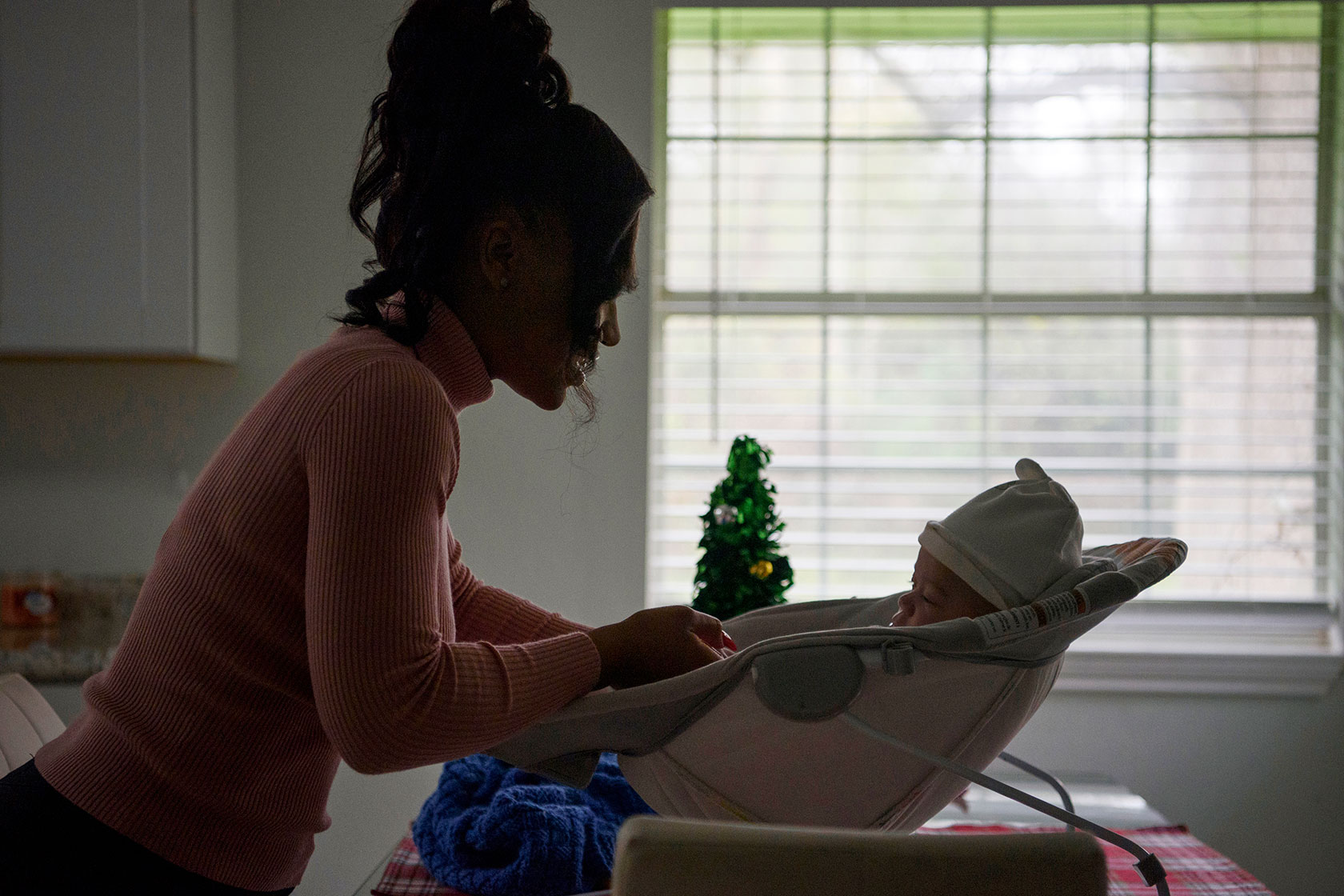 Photo shows a silhouette of a woman looking at her baby, who lies in a baby holder