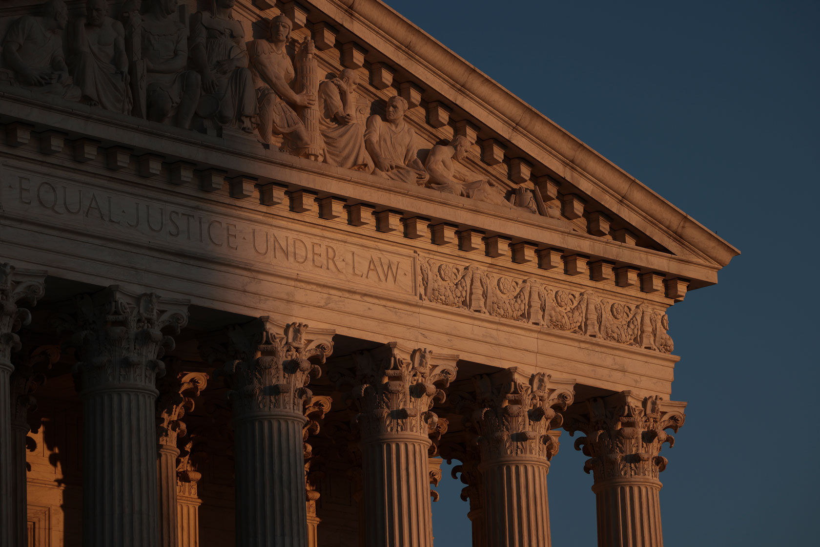Photo shows a close-up shot of the top of the U.S. Supreme Court building with the sunset light hitting it, against a blue sky.