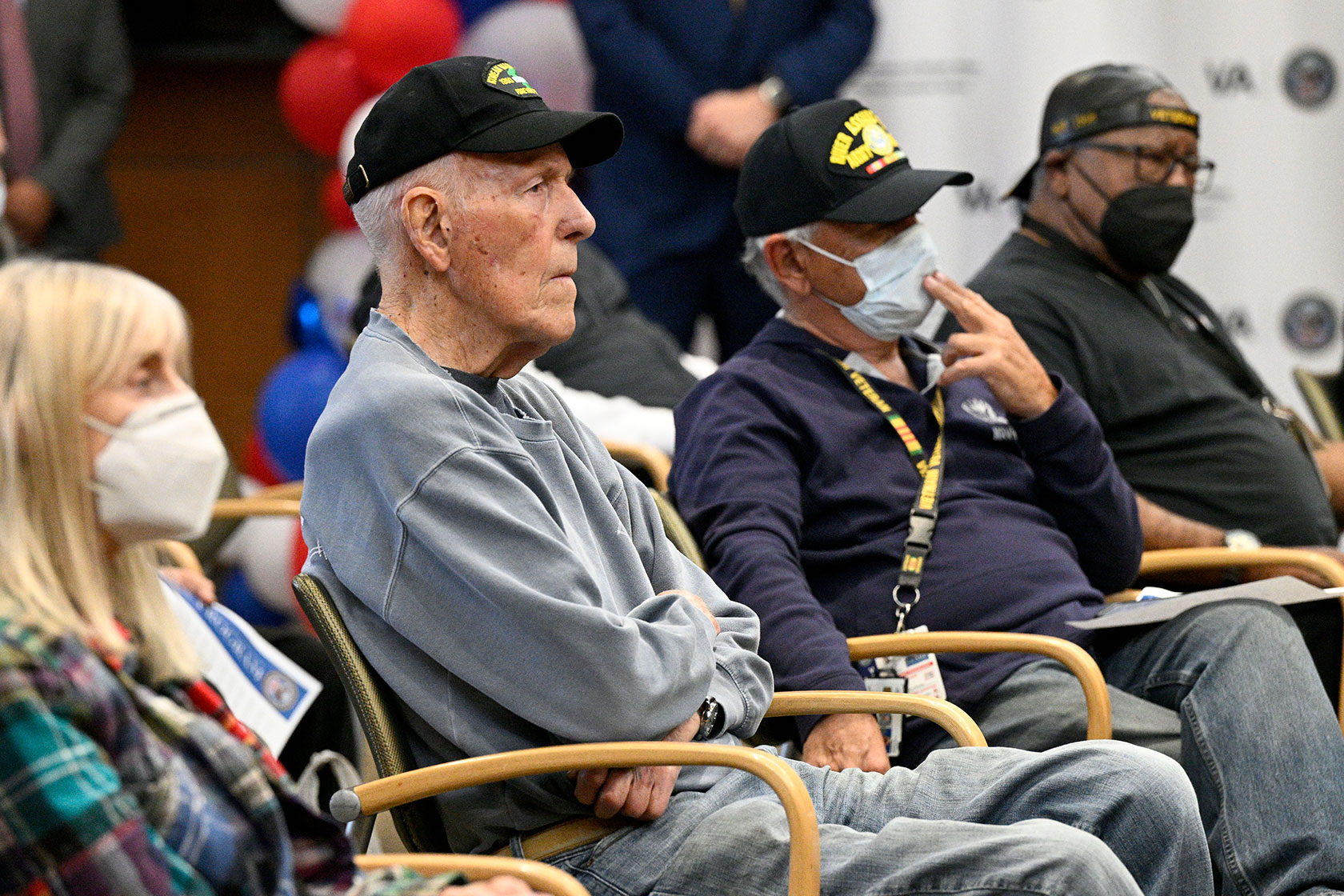 Photo shows four people sitting in chairs. Two wear masks and three wear black baseball caps.