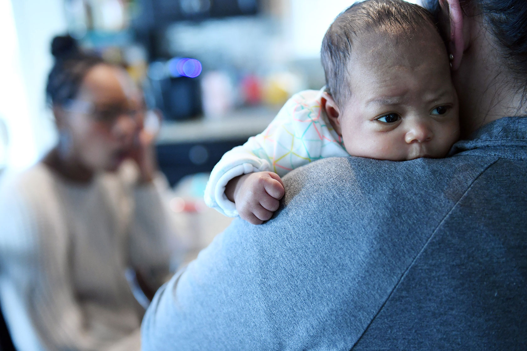 Photo shows a young baby over the shoulder of a woman, with another woman in the background