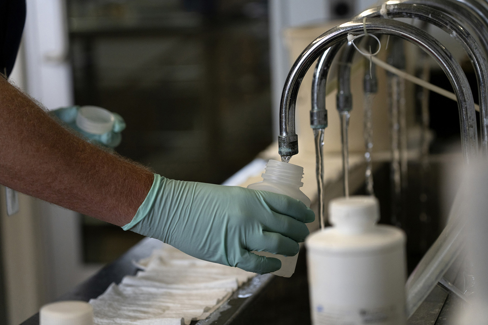 Hand holding collection jug under one faucet in a line of running faucets