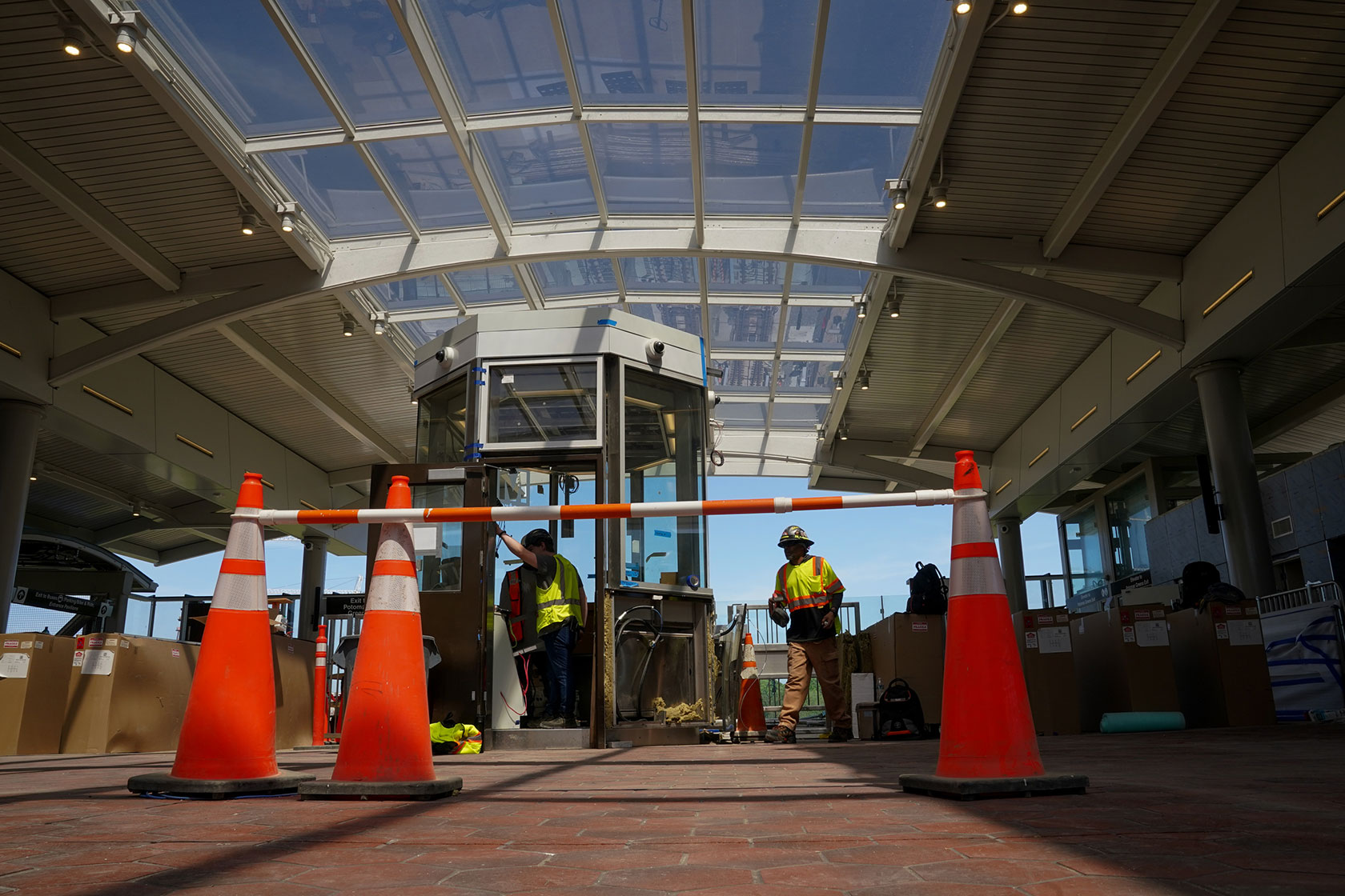 Photo shows two construction workers in bright clothing behind three orange cones at a metro station with glass ceilings