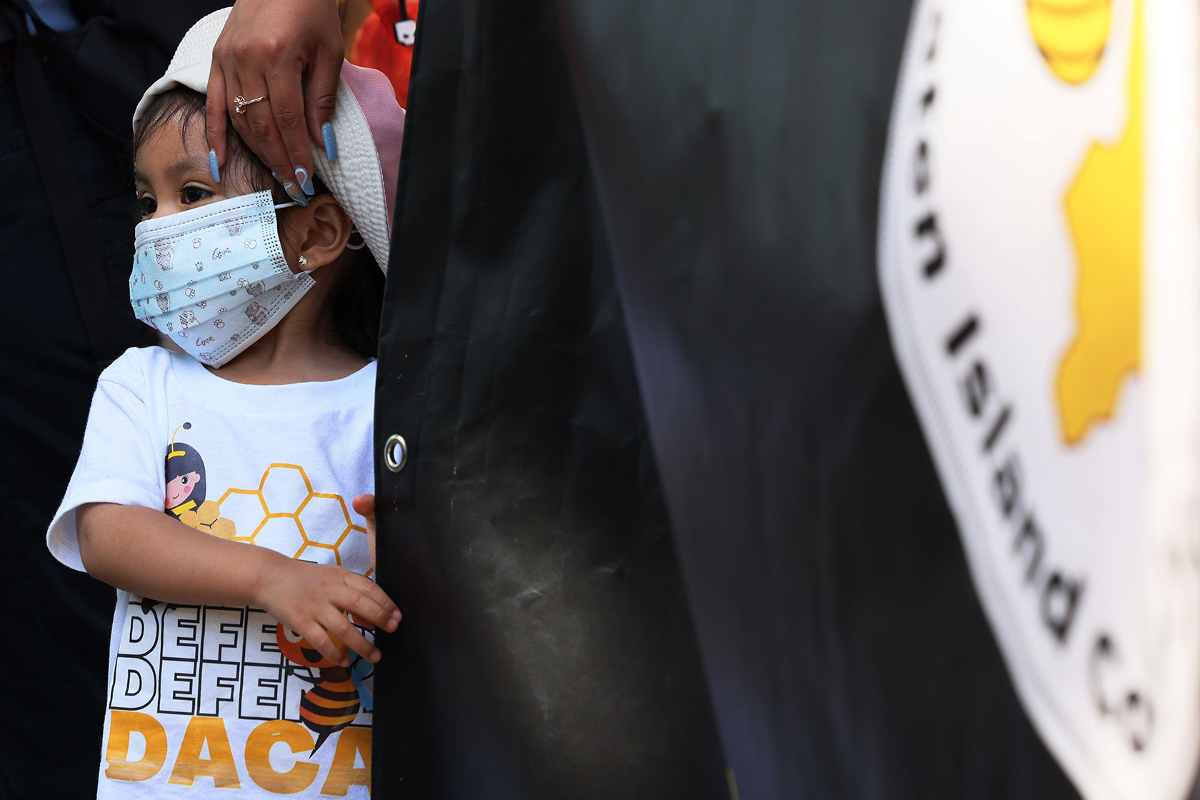 Photo shows a young child wearing a white and yellow shirt and a mask with an adult's hand gently touching her head