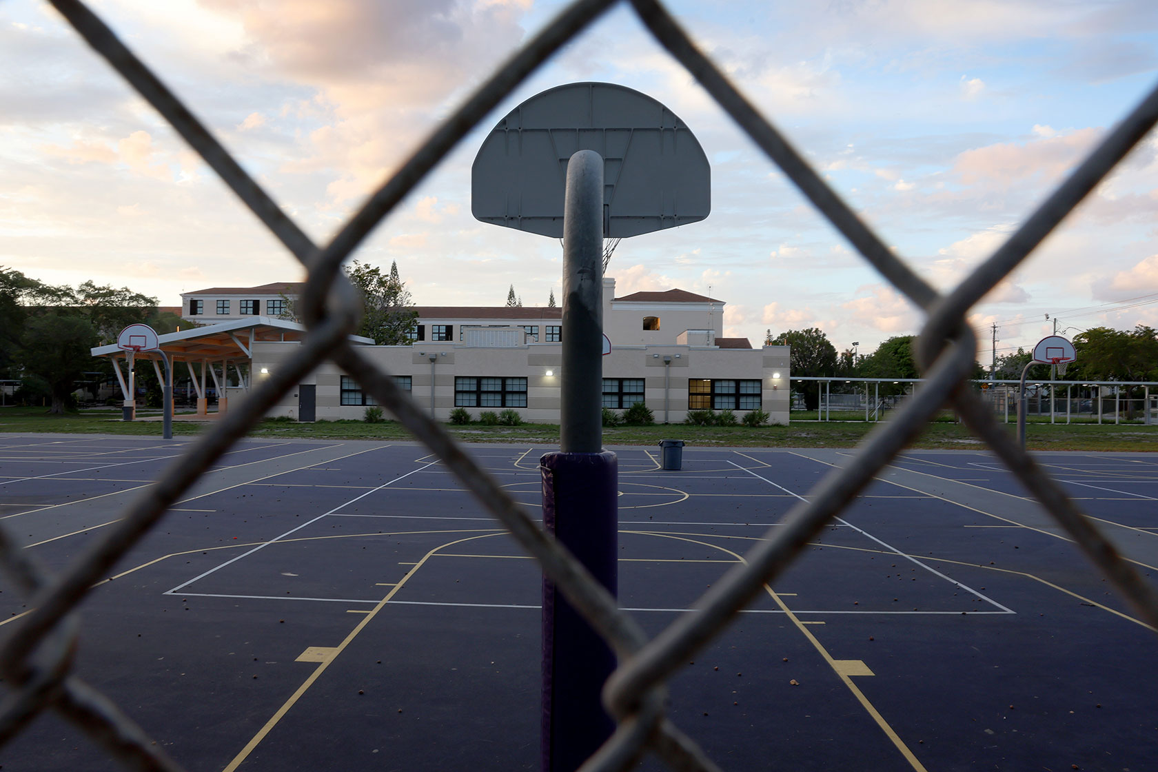 Photo shows an empty basketball court facing a one-story white school, with a chainlink fence between the camera lens and the basketball court