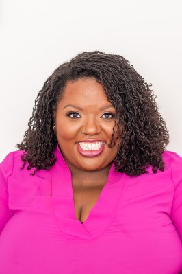 Photo shows Raven Freeborn smiling for a headshot against a white background, wearing a bright pink top