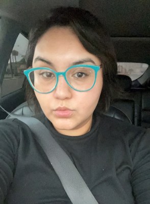 Photo shows Andrea Contreras wearing a black shirt and turquoise glasses taking a selfie, as she sits in a car with a seat belt on