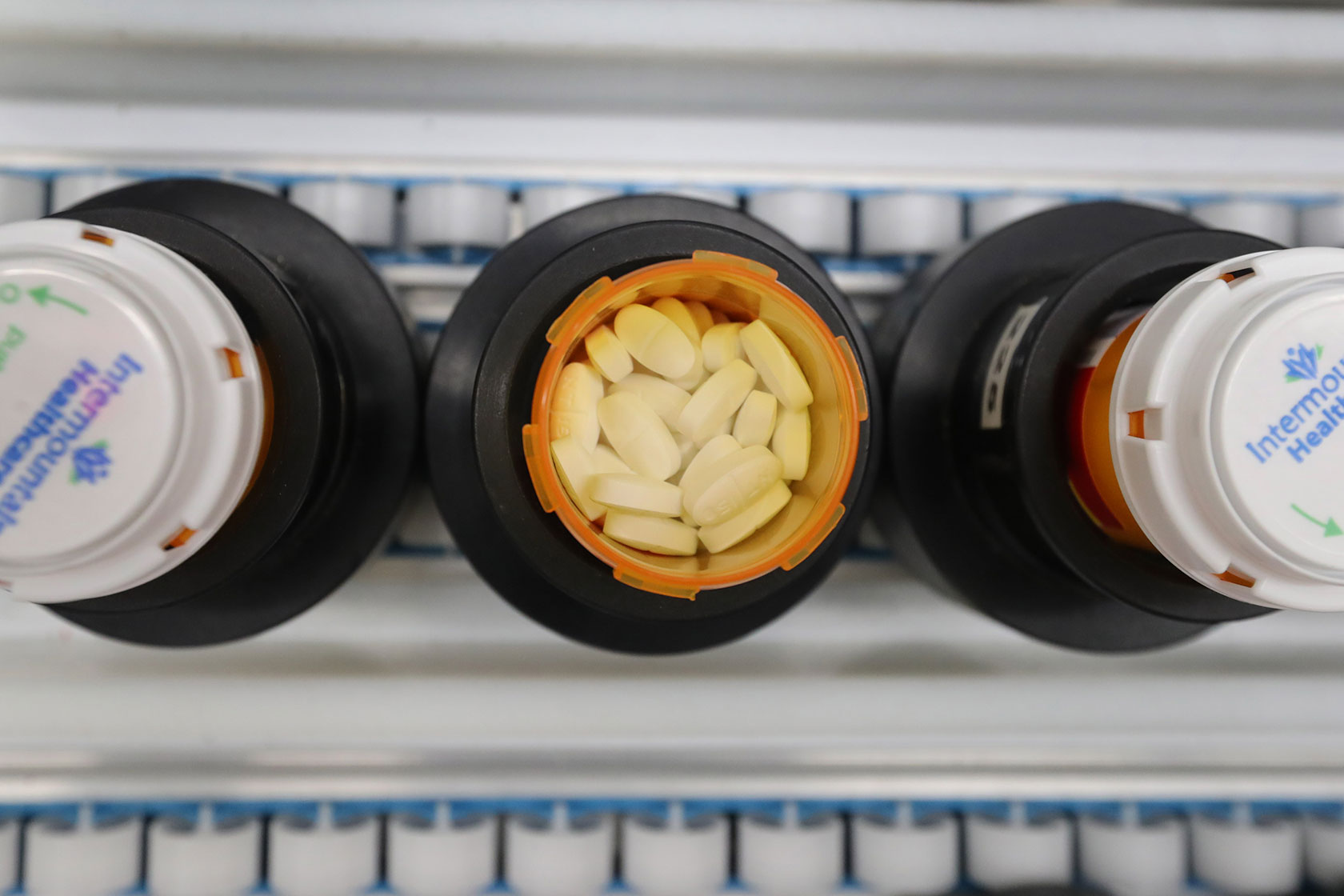 Photo shows three orange prescription drug bottles—one full with the cap off, and two next to it with the caps screwed on—on an assembly line