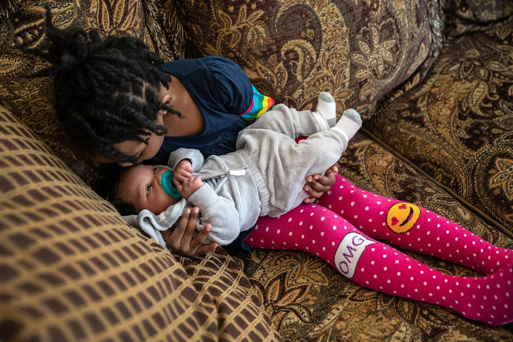 Photo shows a girl in pink stockings holding her baby brother and kissing his face on a multipatterned couch