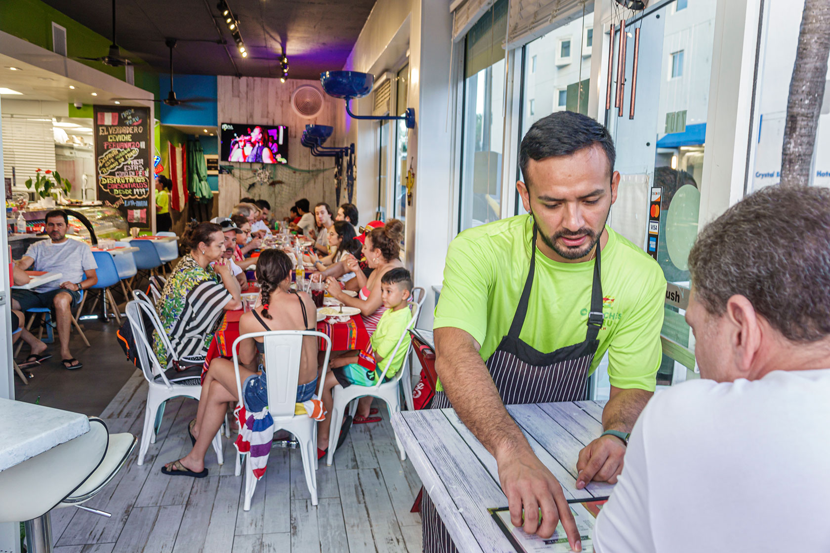 Photo shows a server wearing a bright green shirt and an apron pointing to items on a menu in front of a customer