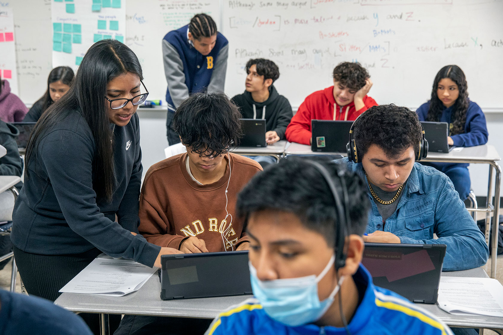 Photo shows two students in a classroom standing up as they help sitting students on their laptops