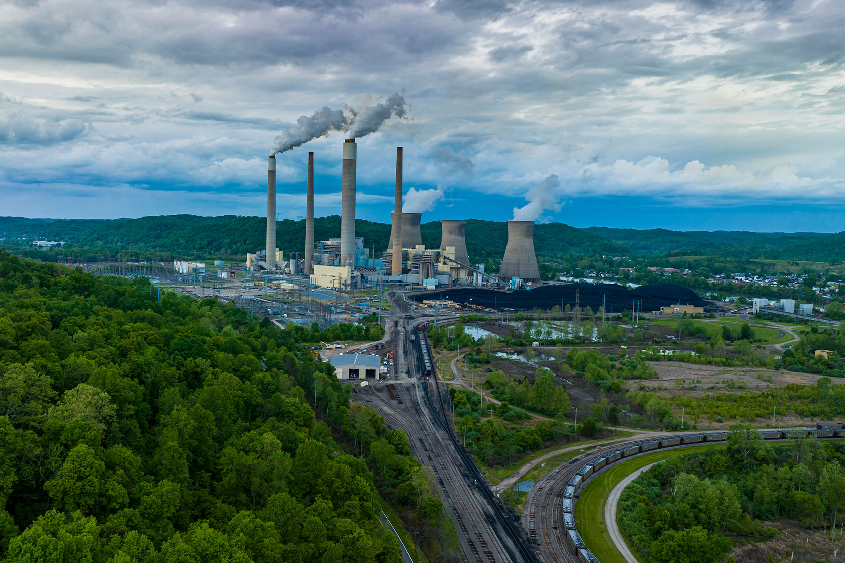 Photo shows smoke rising out of smokestacks at a power plant nestled among green hills against a cloudy sky