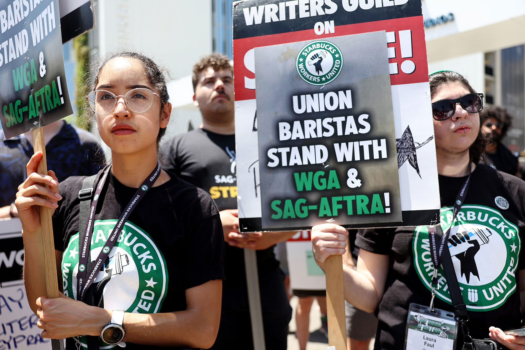 Photo shows a young woman wearing a black Starbucks Workers United shirt, and other Starbucks employees, holding signs supporting the SAG-AFTRA and Writers Guild of America striking workers
