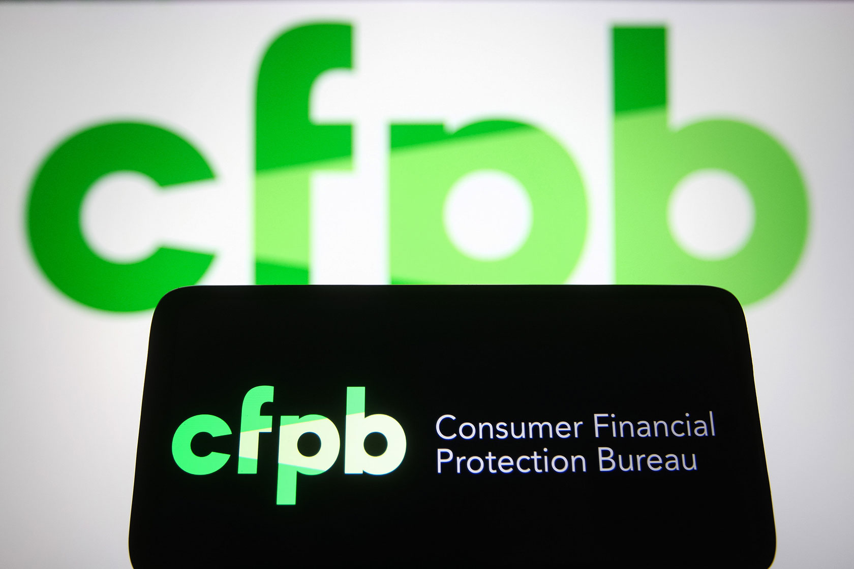 Photo shows the CFPB lime-green logo against a black background on a smartphone, with a larger white screen displaying the green CFPB logo in the background