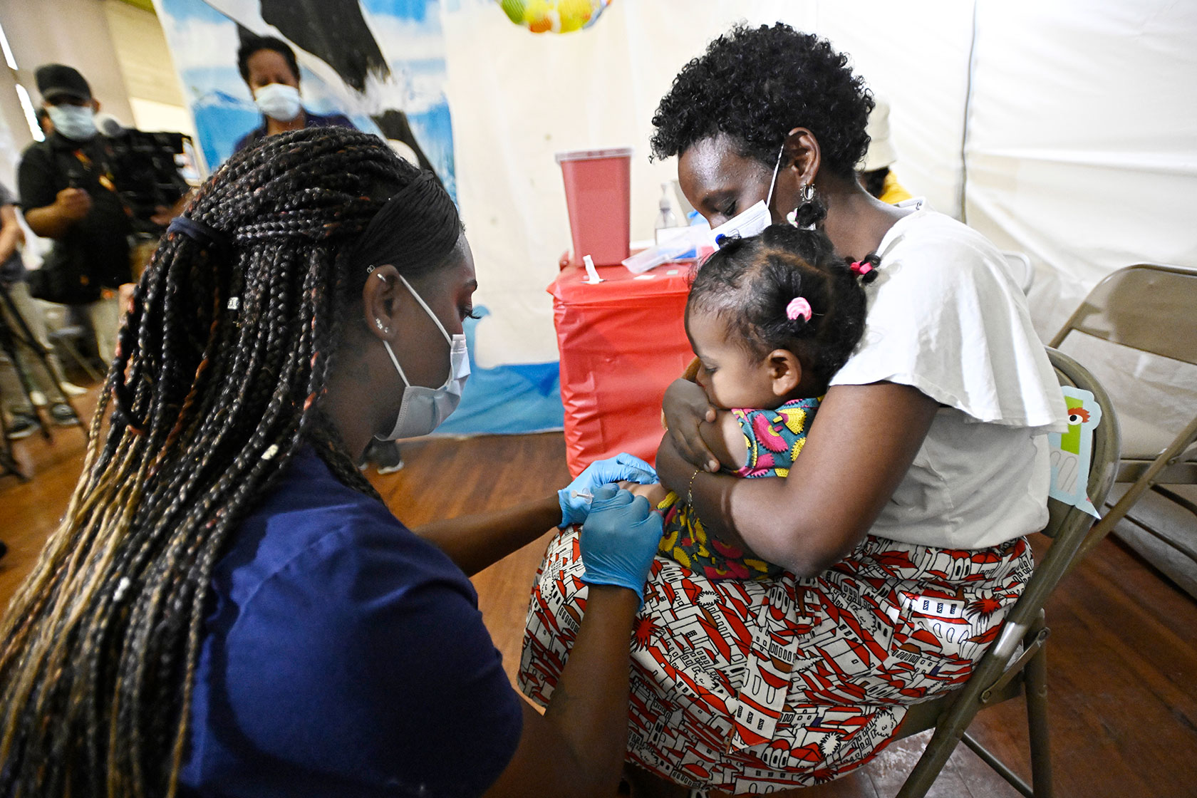 Photo shows a mother wearing a mask with her 1-year-old daughter on her lap. A nurse in dark-blue scrubs gives the child a vaccine in her leg
