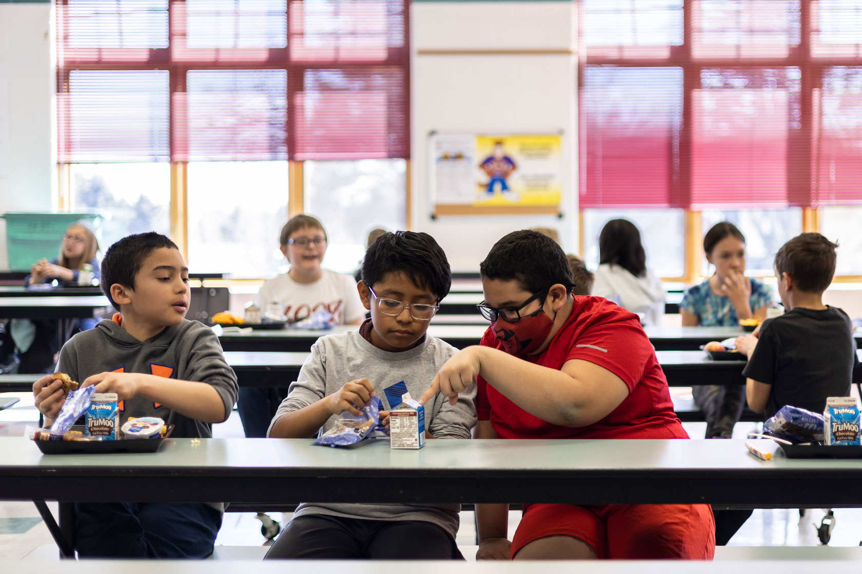 Three elementary students sit and compare lunches at a cafeteria table.