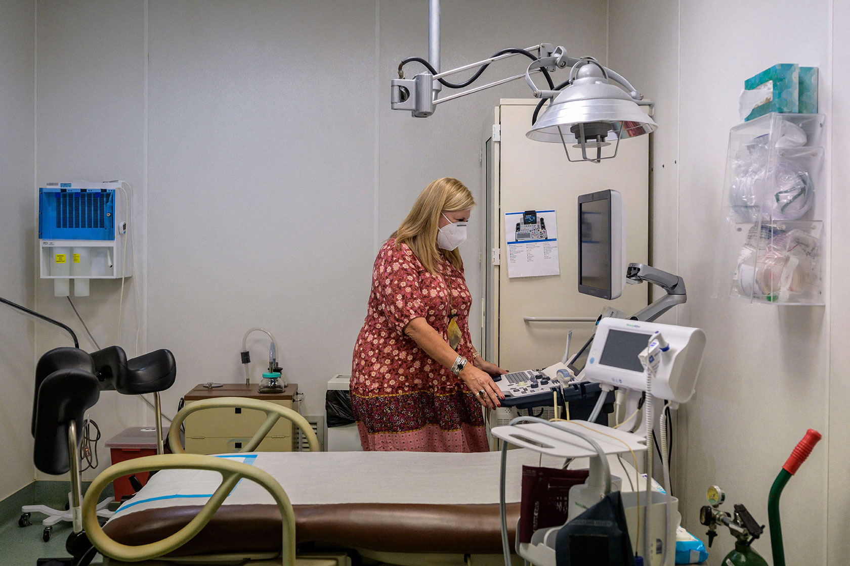 Photo shows a woman wearing a mask standing next to a gurney in an exam room