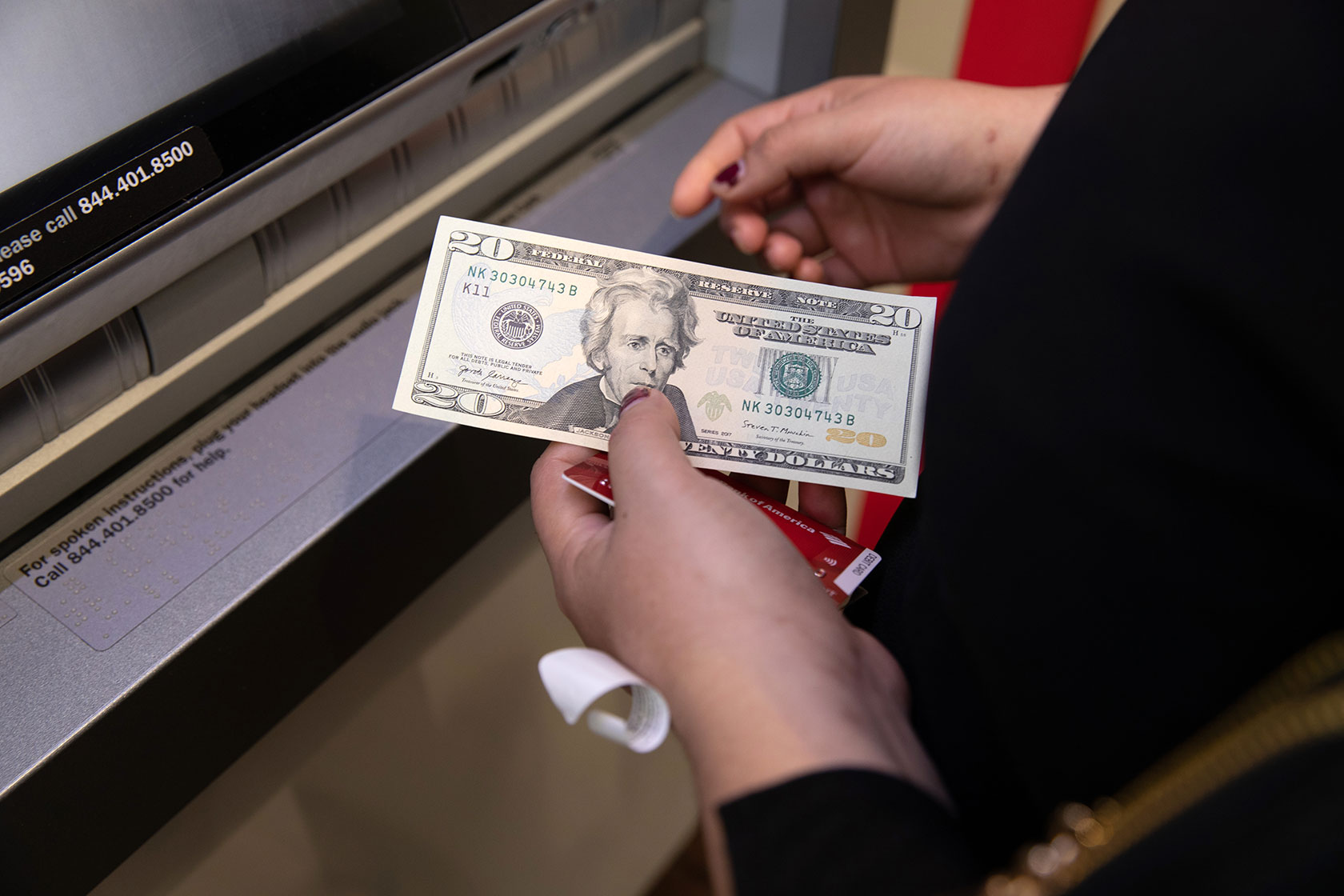 Photo shows a closeup of a person's hands in front of an ATM. In one hand, the person holds a $20 bill