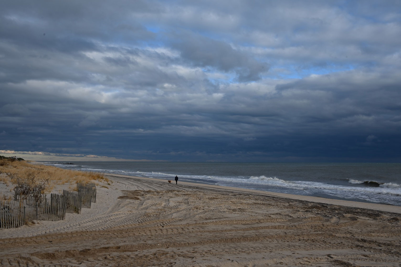Photo shows a man and his dog in the distance on an otherwise empty beach on a cloudy day