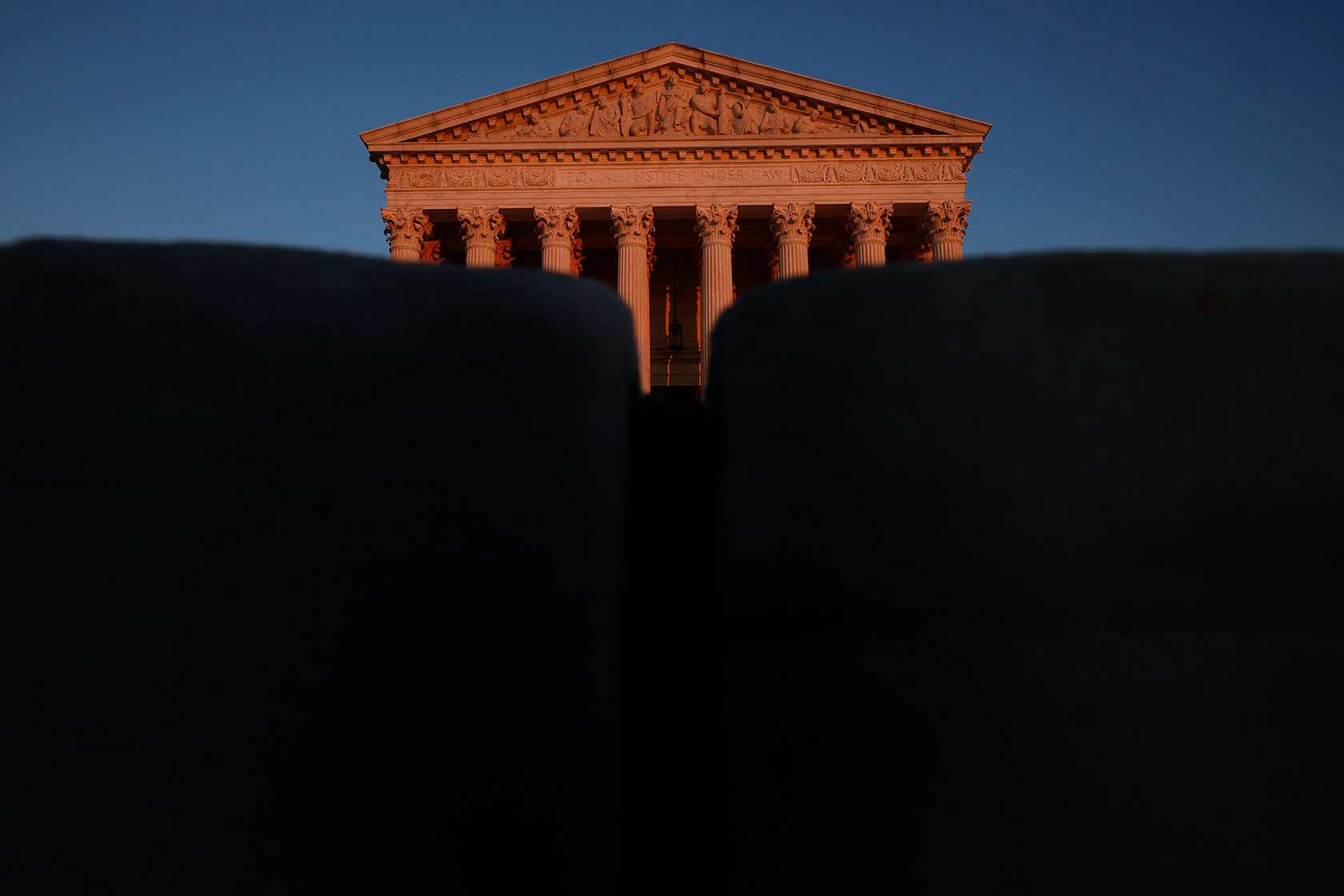Photo shows a partially obscured view of the top of the Supreme Court building reflecting orange sunlight against a deep blue sky