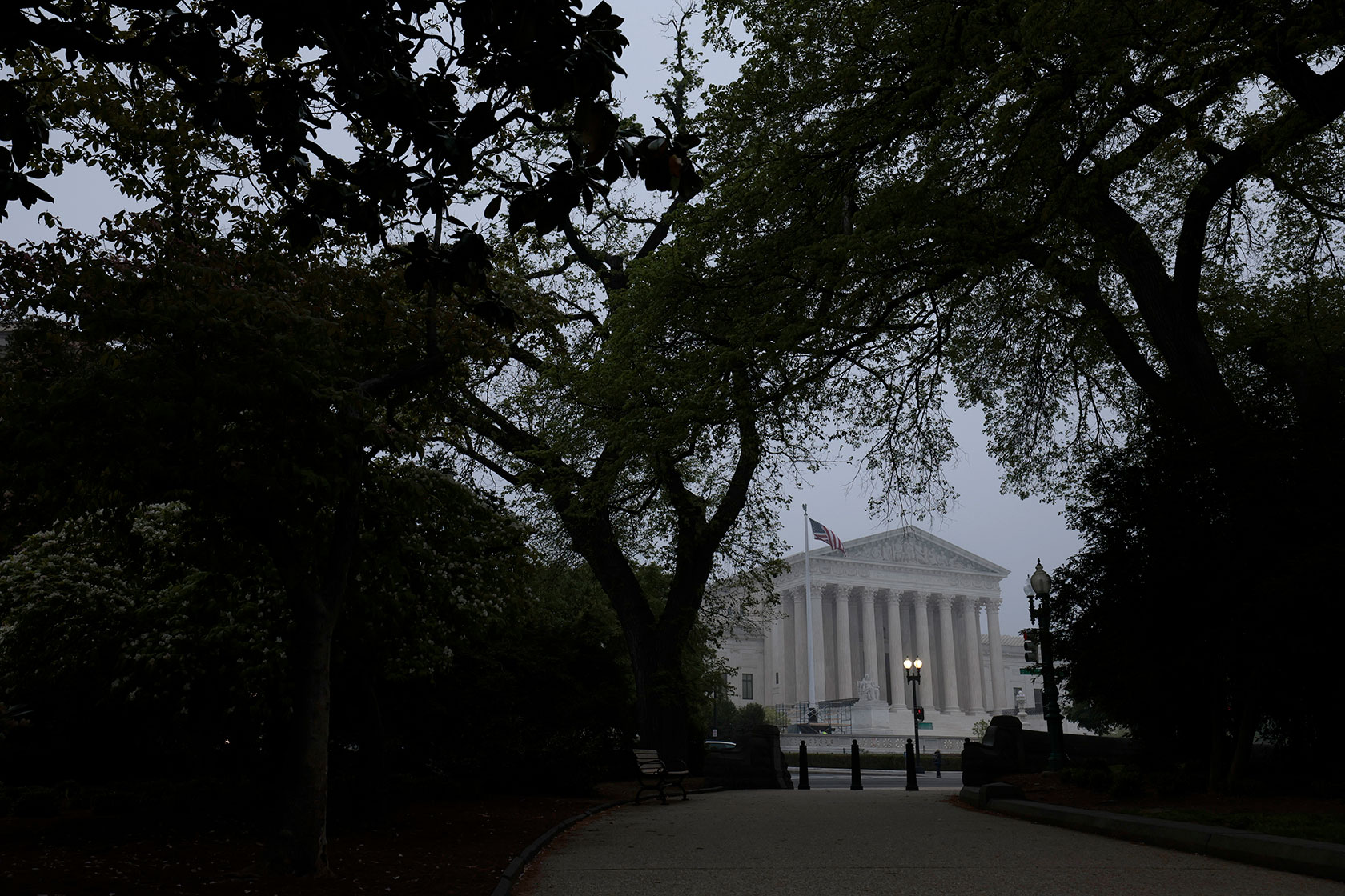 Photo shows the Supreme Court building with an American flag waving in front, through a view of silhouetted trees against a grey cloudy sky