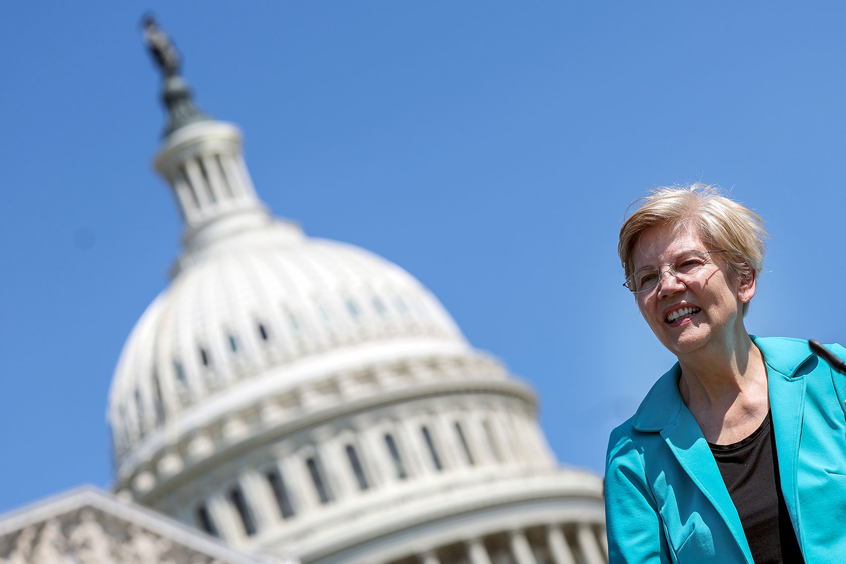 Photo shows a tilted image of Elizabeth Warren wearing turquoise as she speaks in front of the U.S. Capitol building dome against a blue sky