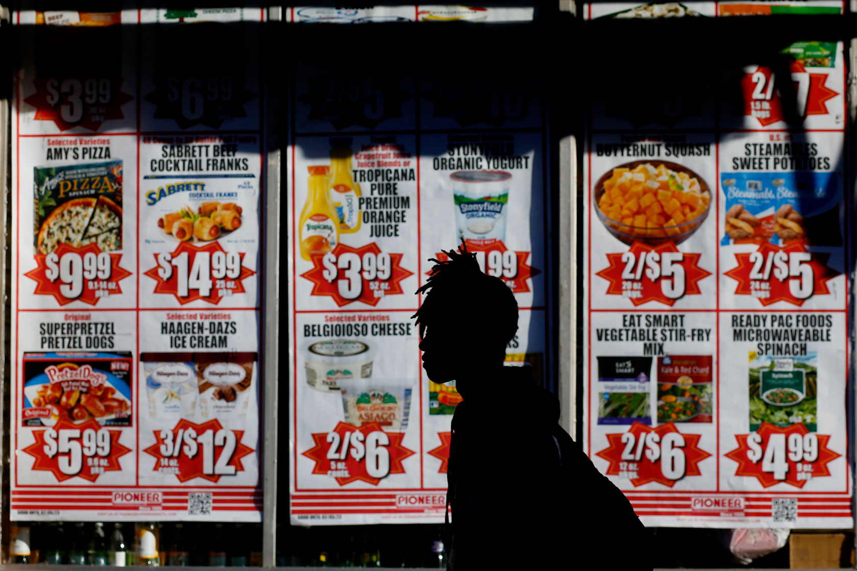 Photo shows a silhouette of a person walking past a window display with rows of signs displaying photos of foods and their associated prices