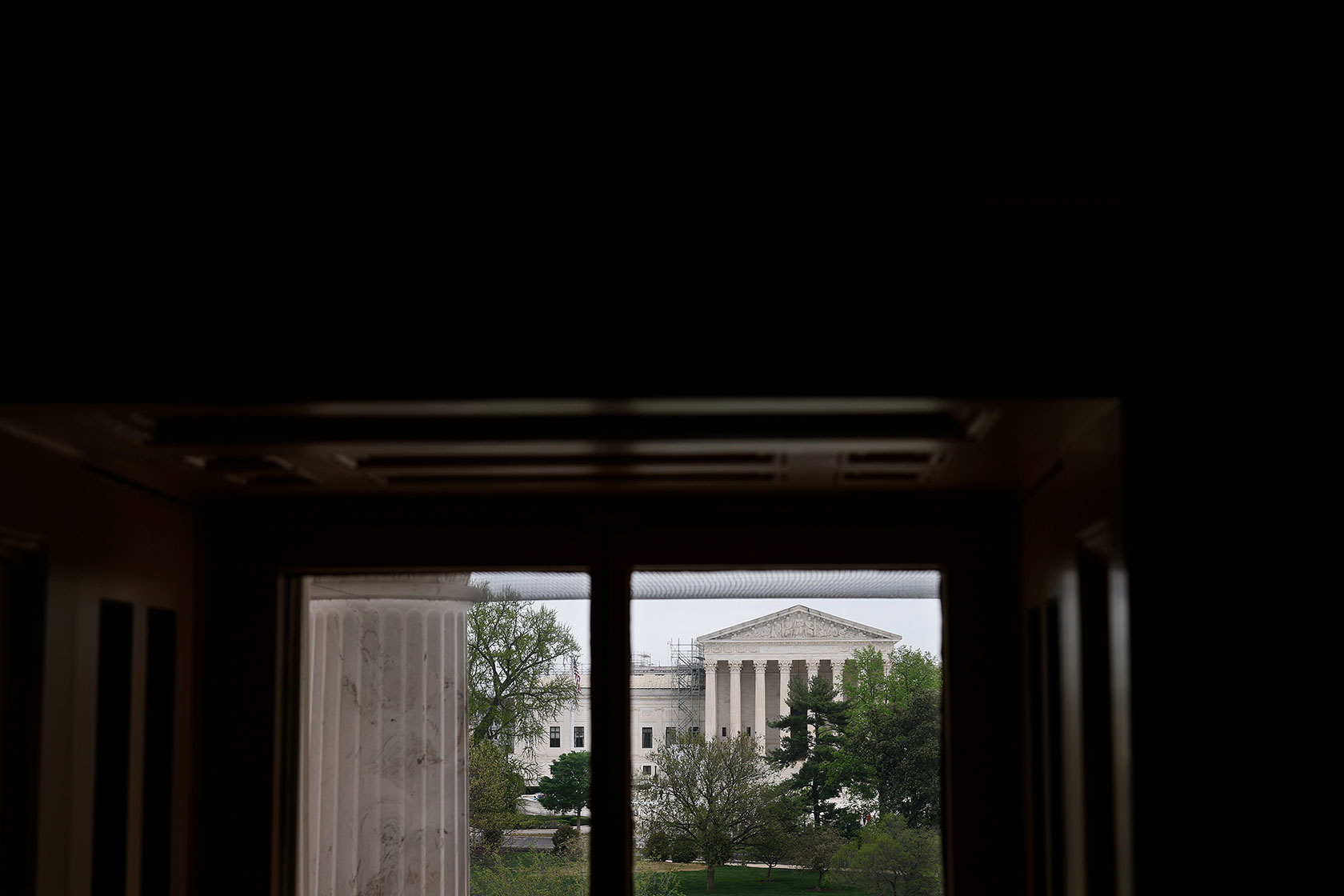 Photo shows a view of the U.S. Supreme Court building against a light grey sky, viewed through a dark window