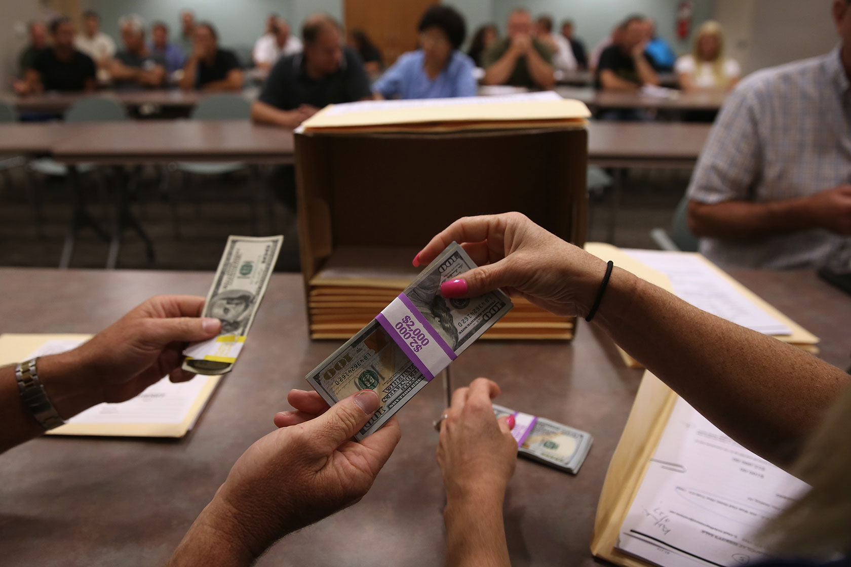 Photo shows two hands exchange bundles of 100 dollar bills in front of rows of people sitting in a courthouse