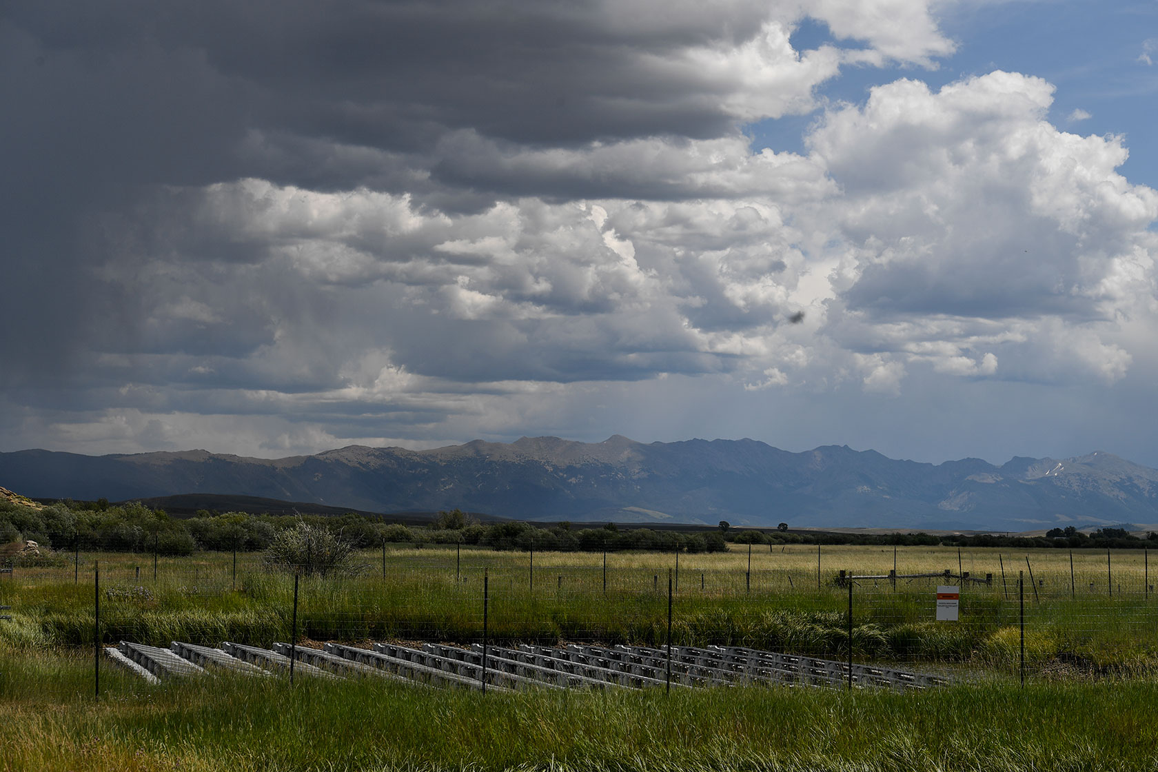Photo shows a solar panel array in a green field with mountains in the background and a partly cloud sky