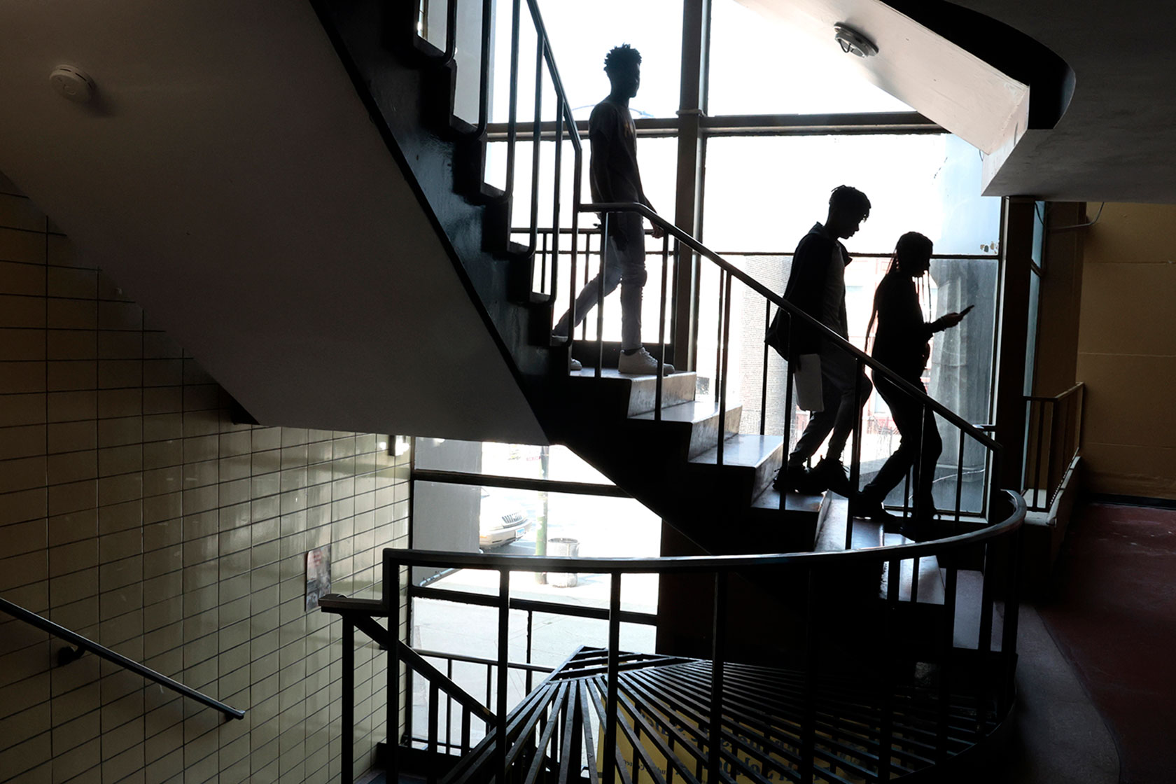Photo shows three students walking down stairs, silhouetted against a large window showing a grey sky outside