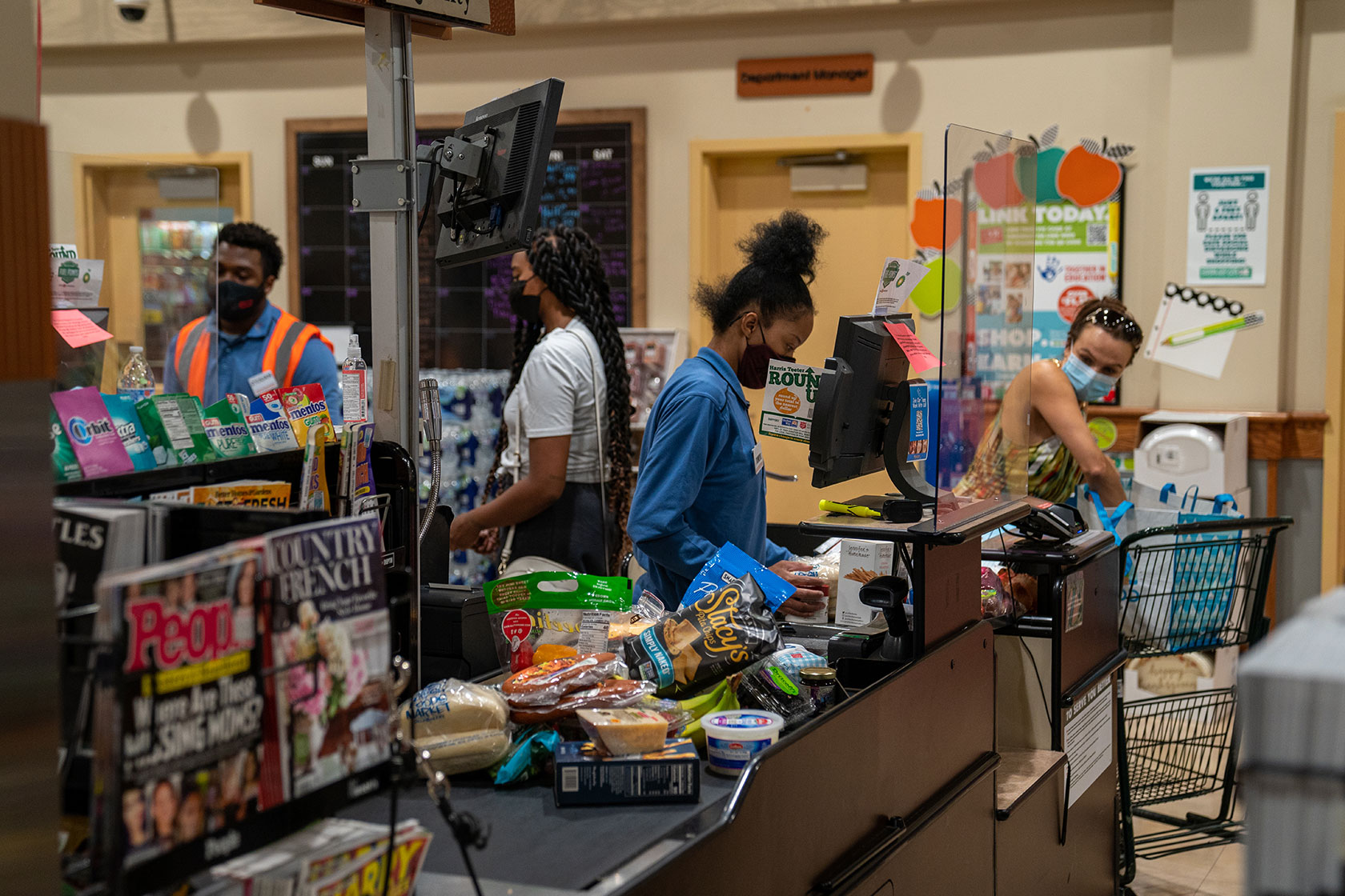 Photo shows a young woman scanning groceries from a full conveyor belt, as the customer helps bag the items.