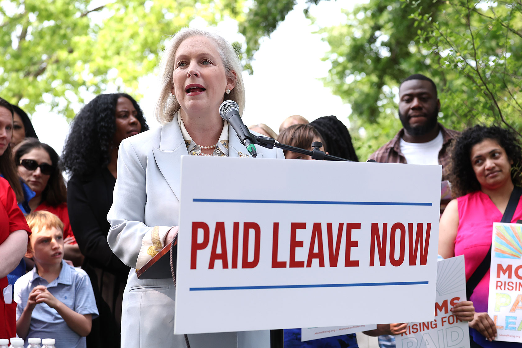 Photo shows Kirsten Gillibrand wearing all white, speaking at a podium with a sign in front of it that says 