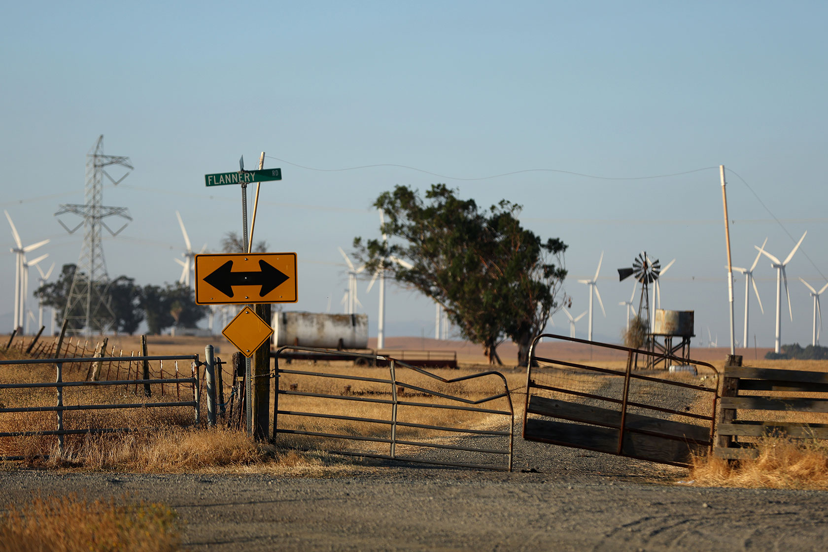 Photo shows a street sign in front of an open field with golden grass, a tree, and white wind turbines against a dusty blue sky