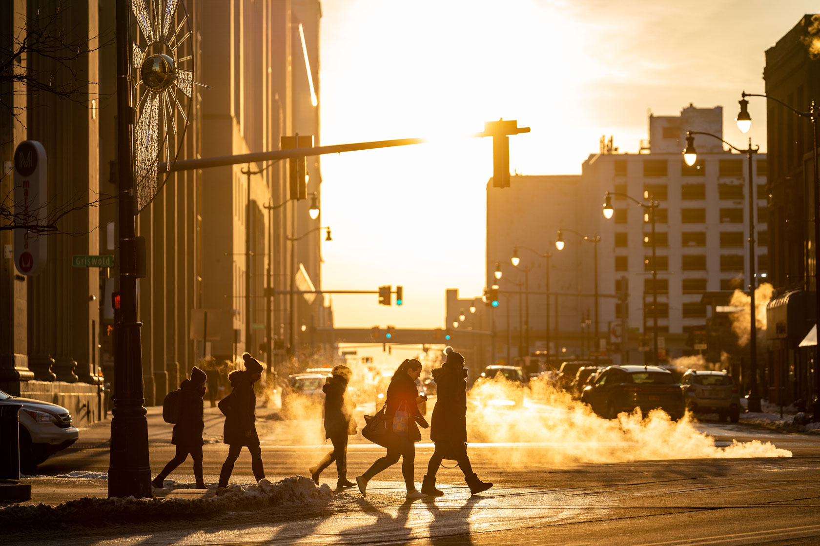 Photo shows people walking across a street as the sun sets in the distance.