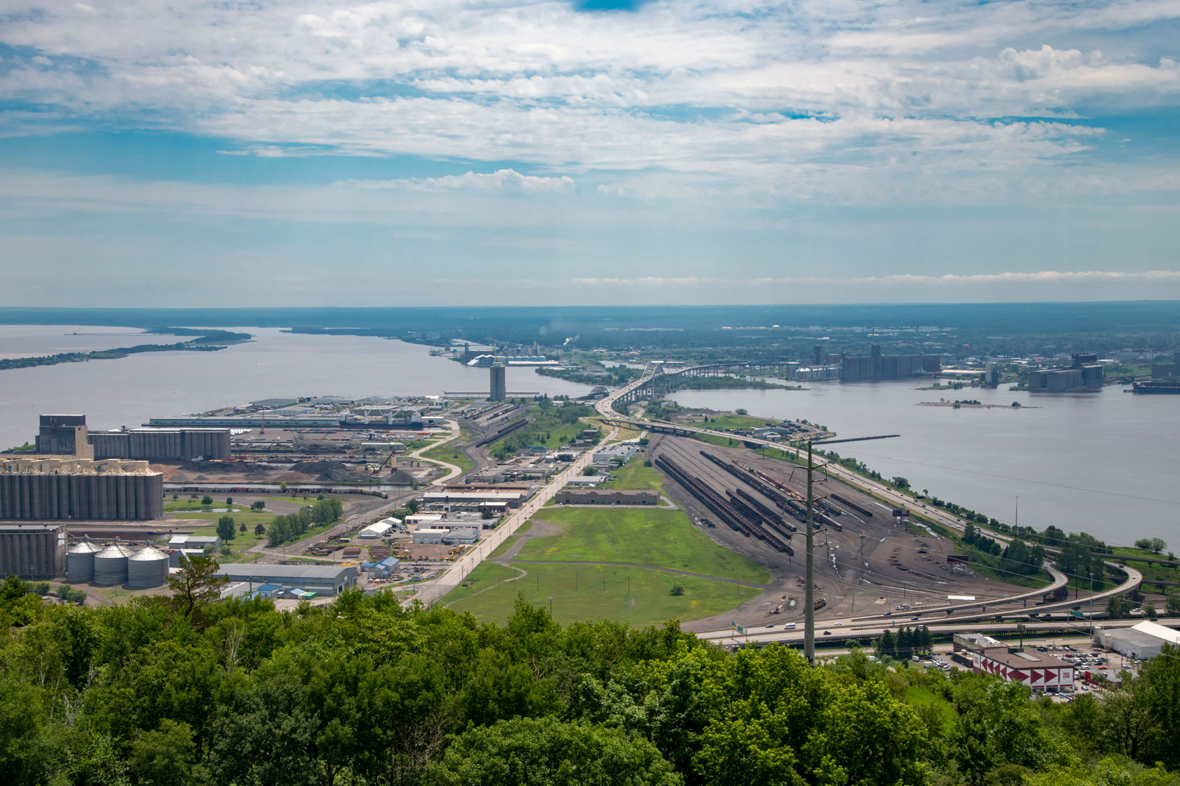 Photo shows an aerial view of the harbor with an expanse of green grass and trees, flanked by bodies of water on both sides