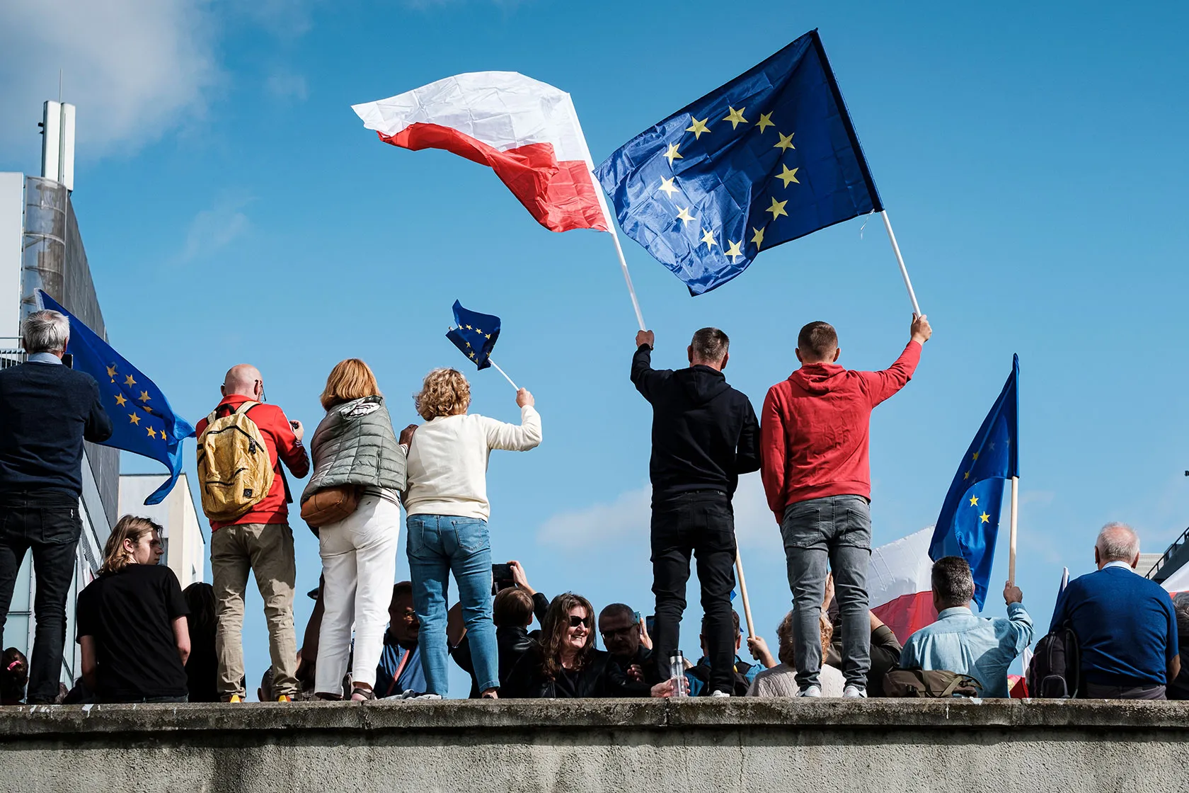 Photo shows five people standing on a wall waving the EU flag and the Polish flag against a clear blue sky