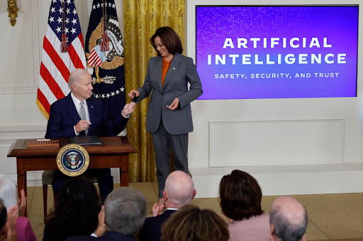 A photo of President Joe Biden and Vice President Harris in the White House signing the executive order on AI. President Joe Biden is seated and hands Vice President Kamala Harris a pen. There is a sign behind them with white text on a purple background reading: 