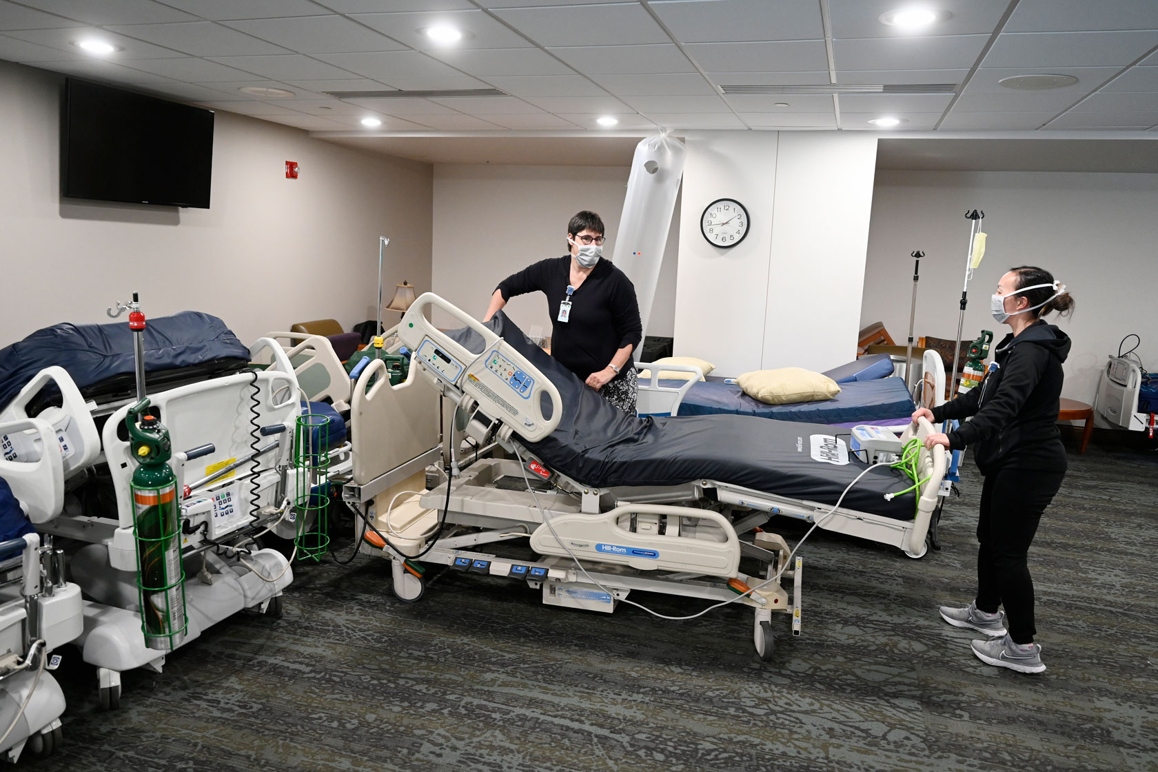 Photo shows two workers moving a hospital bed, with a couple of other beds in the foreground and background.