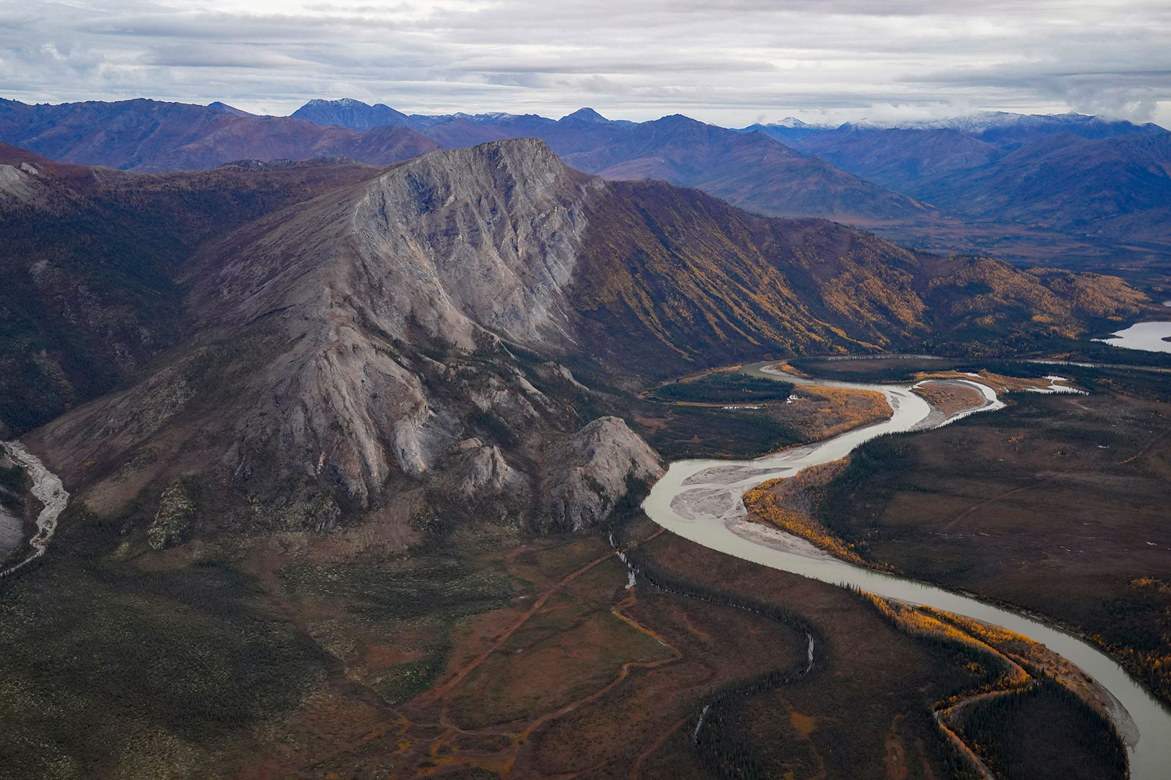 Photo shows an aerial view of a mountain range, mostly bare with no trees, against a grey, cloudy sky. A serpentine river snakes through the valleys between the mountains.