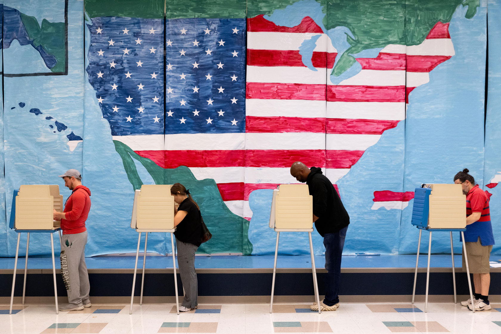 Four voters in a line fill out their ballots in front of a large map of the United States on the wall.