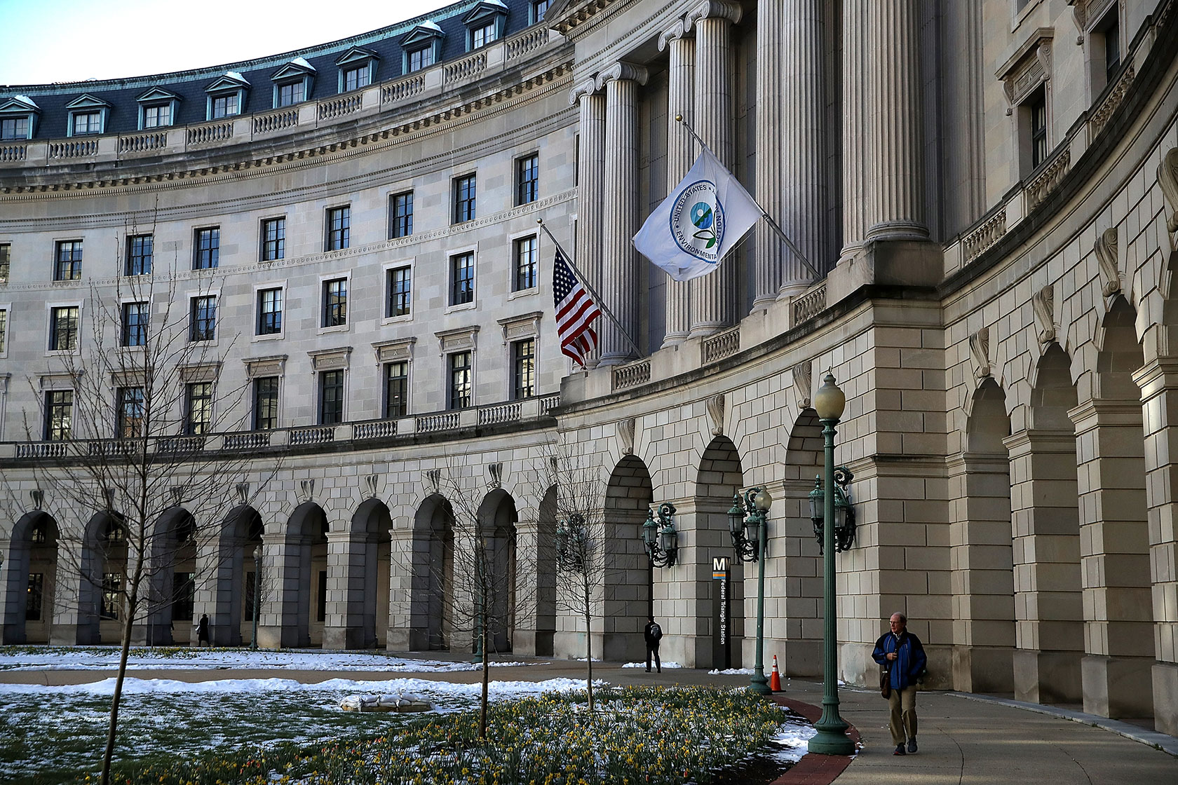Photo shows a partial view of the EPA building, with a U.S. and EPA flag flying in front, and two people walking on the sidewalk