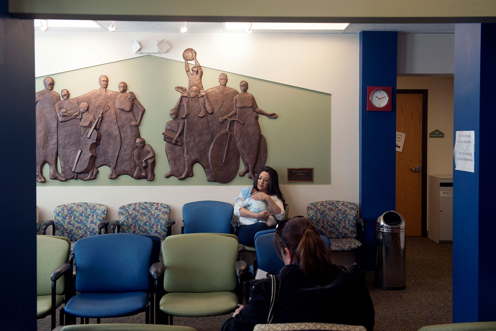 Photo shows a woman holding her young baby in a waiting room, with a mural in the background