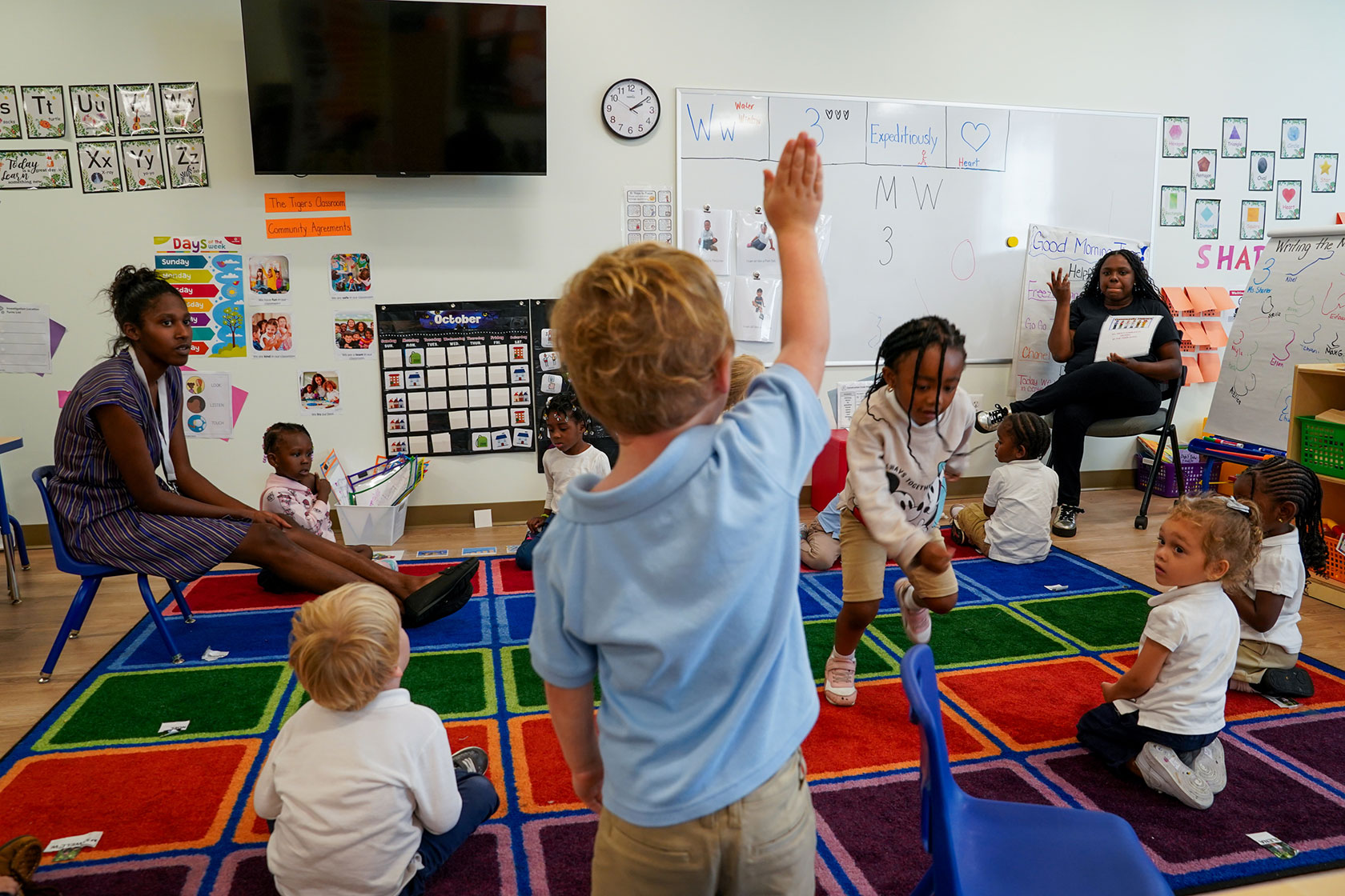 A preschool student raises their hand as teachers lead a class at an early learning public charter school.